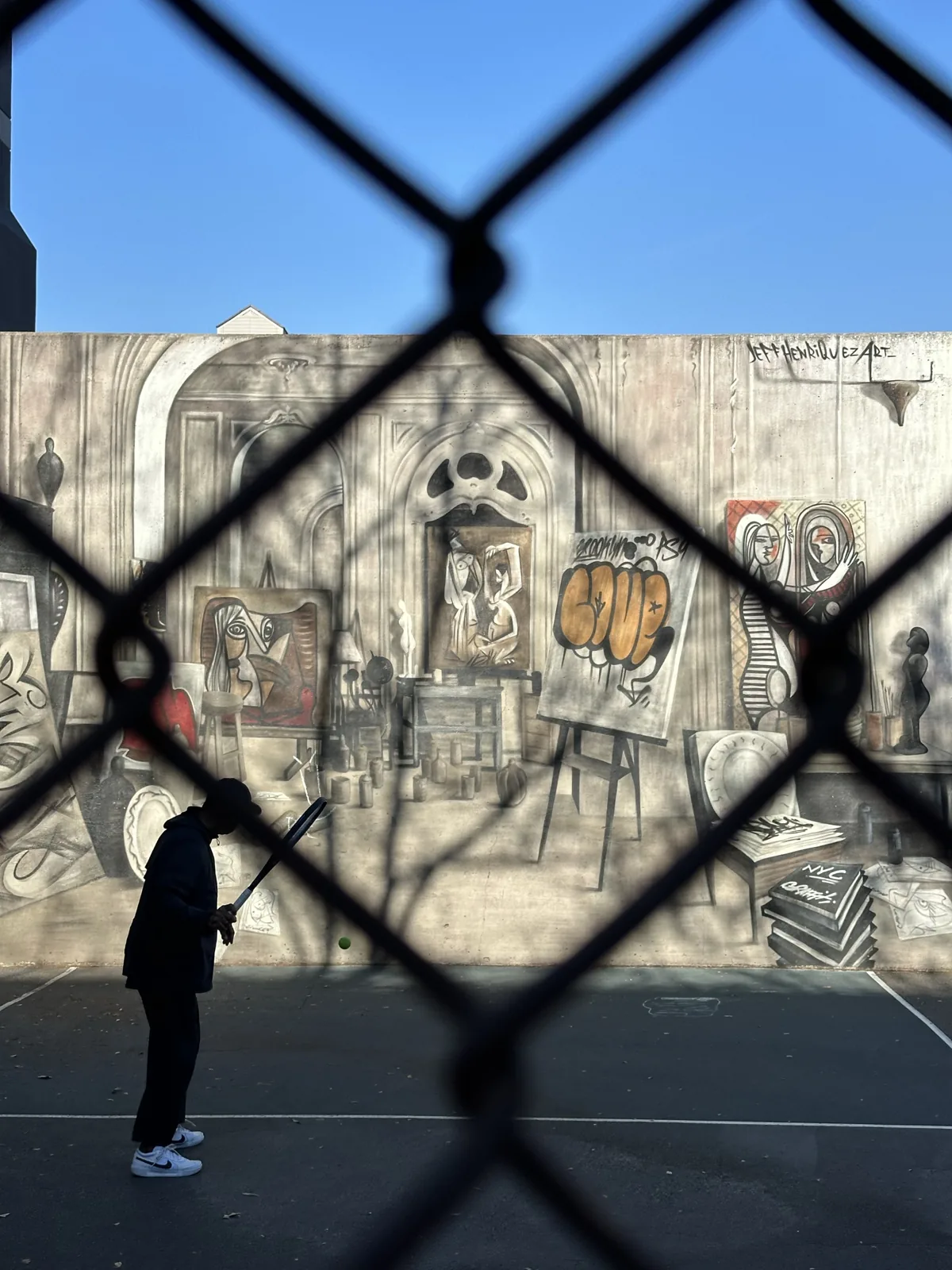A person holding a bat on a mural-lined court, seen through a chain-link fence.